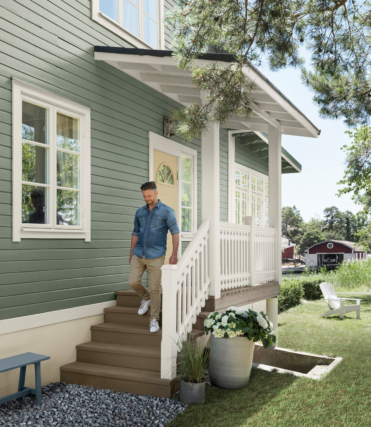 Man klädd i jeansskjorta som går nerför trappan vid grönt hus målad i Jotun 7653 Stockholmsgrön med vit veranda, omgiven av grönskande trädgård och utomhusstolar