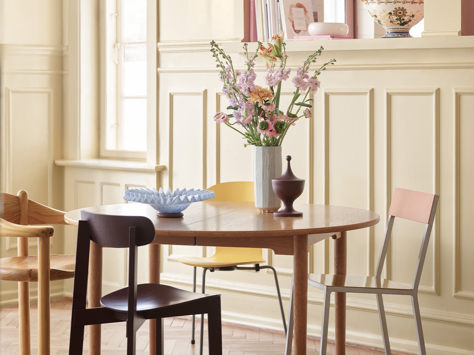 Elegant spiseplass med rundt trebord, ulike designstoler og vase med blomster i lyst, klassisk interiør