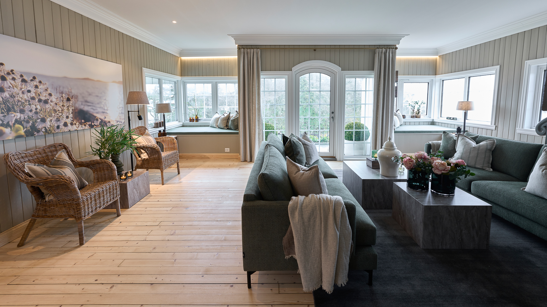 Lys og elegant stue med store vinduer, grønne sofaer, og rottingstoler. Rommet har moderne innredning med planter og kunstverk, samt et rundt sofabord med blomsterdekorasjon, beige vegger malt i Jotun 1939 Sandbeige