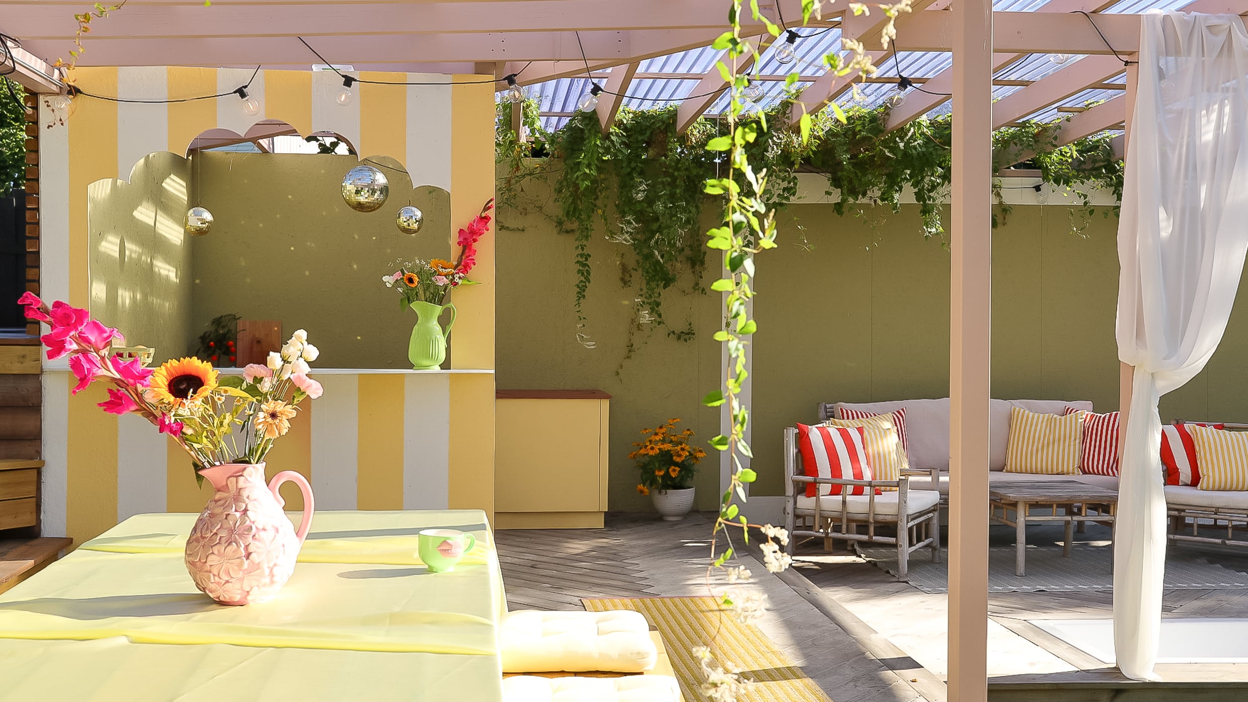 Utekjøkken på terrasse med rosa tak og stolper malt i Jotun Mellomrosa, med bar malt i gul og hvitstripet med Jotun Demigul og Jotun Bomull, grønn bakvegg i Jotun Grønnland, tropisk stemning med friske farger og blomster.