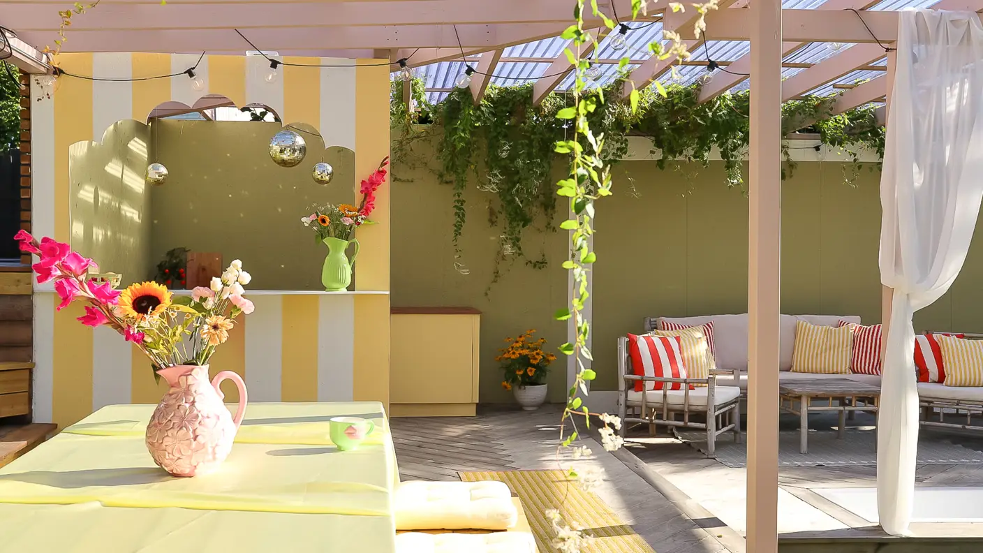 Utekjøkken på terrasse med rosa tak og stolper malt i Jotun Mellomrosa, med bar malt i gul og hvitstripet med Jotun Demigul og Jotun Bomull, grønn bakvegg i Jotun Grønnland, tropisk stemning med friske farger og blomster.