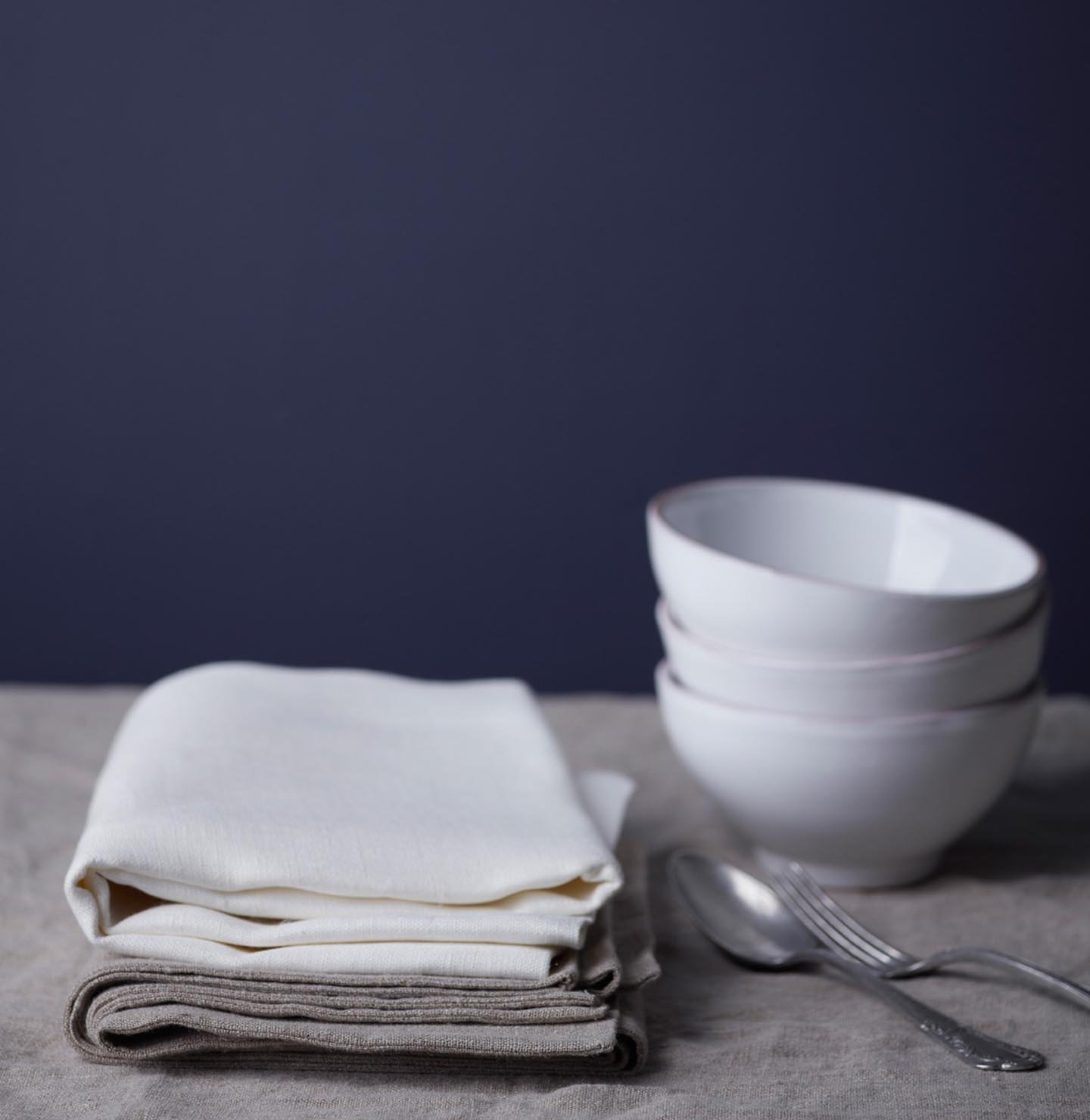 En rödblå vägg, målad med färgen Jotun 4143 Anemon, skapar en lugn bakgrund för en stillsam dukning med vikta linneservetter, staplade vita skålar och enkelt silverbestick.