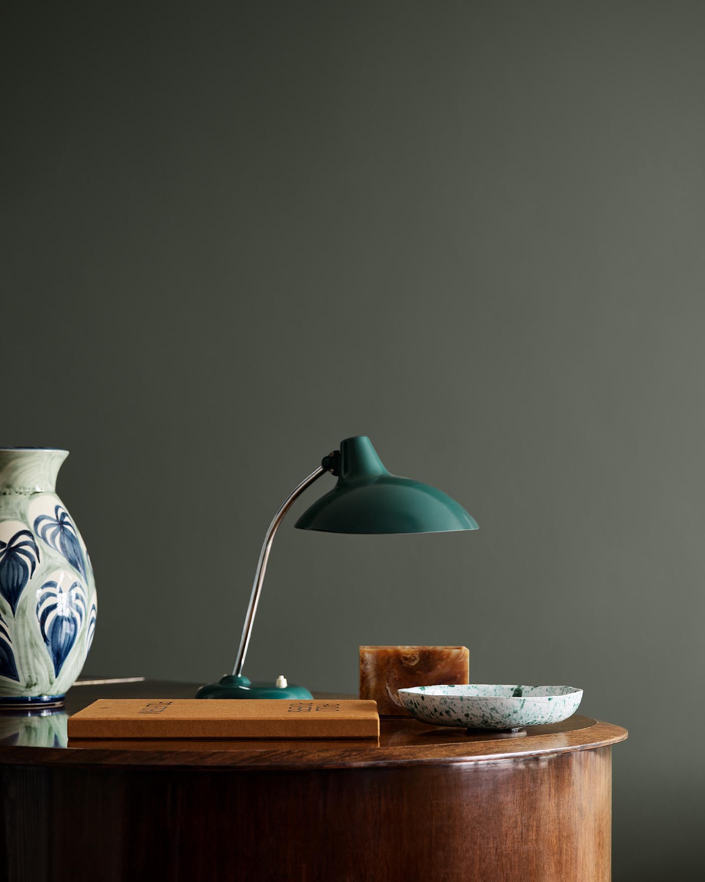 Wooden table with ceramic vase, lamp, and decor against a dark, muted green wall painted in the colour Jotun 7613 Northern Mystic.