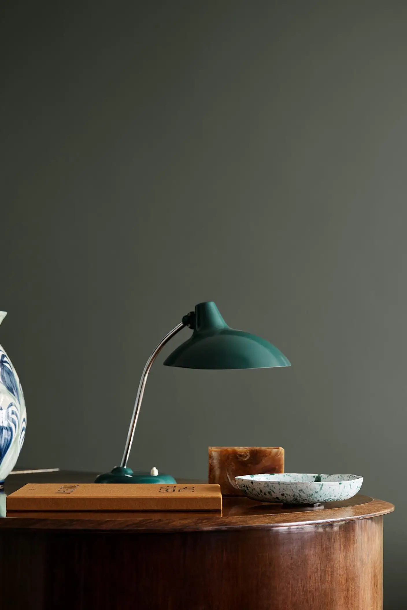 Wooden table with ceramic vase, lamp, and decor against a dark, muted green wall painted in the colour Jotun 7613 Northern Mystic.
