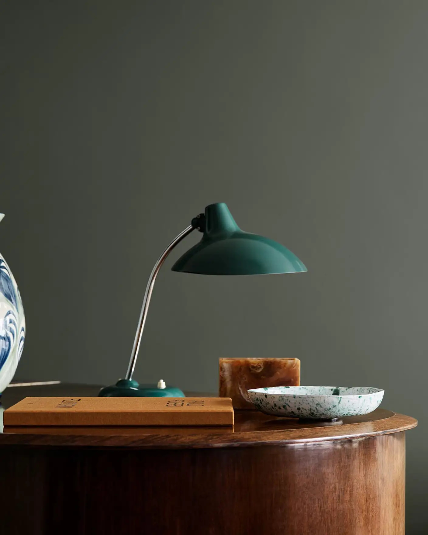 Wooden table with ceramic vase, lamp, and decor against a dark, muted green wall painted in the colour Jotun 7613 Northern Mystic.