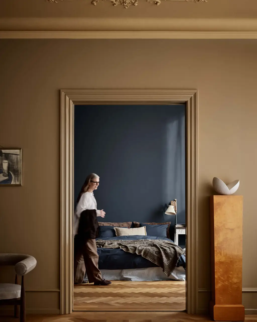 Utsikt gjennom en stuedøråpning i fargen Jotun 1909 Kalkgull og inn i et soverom i Jotun 4403 Midnight Blue, hvor en person går forbi en seng med mønstret sengetøy.