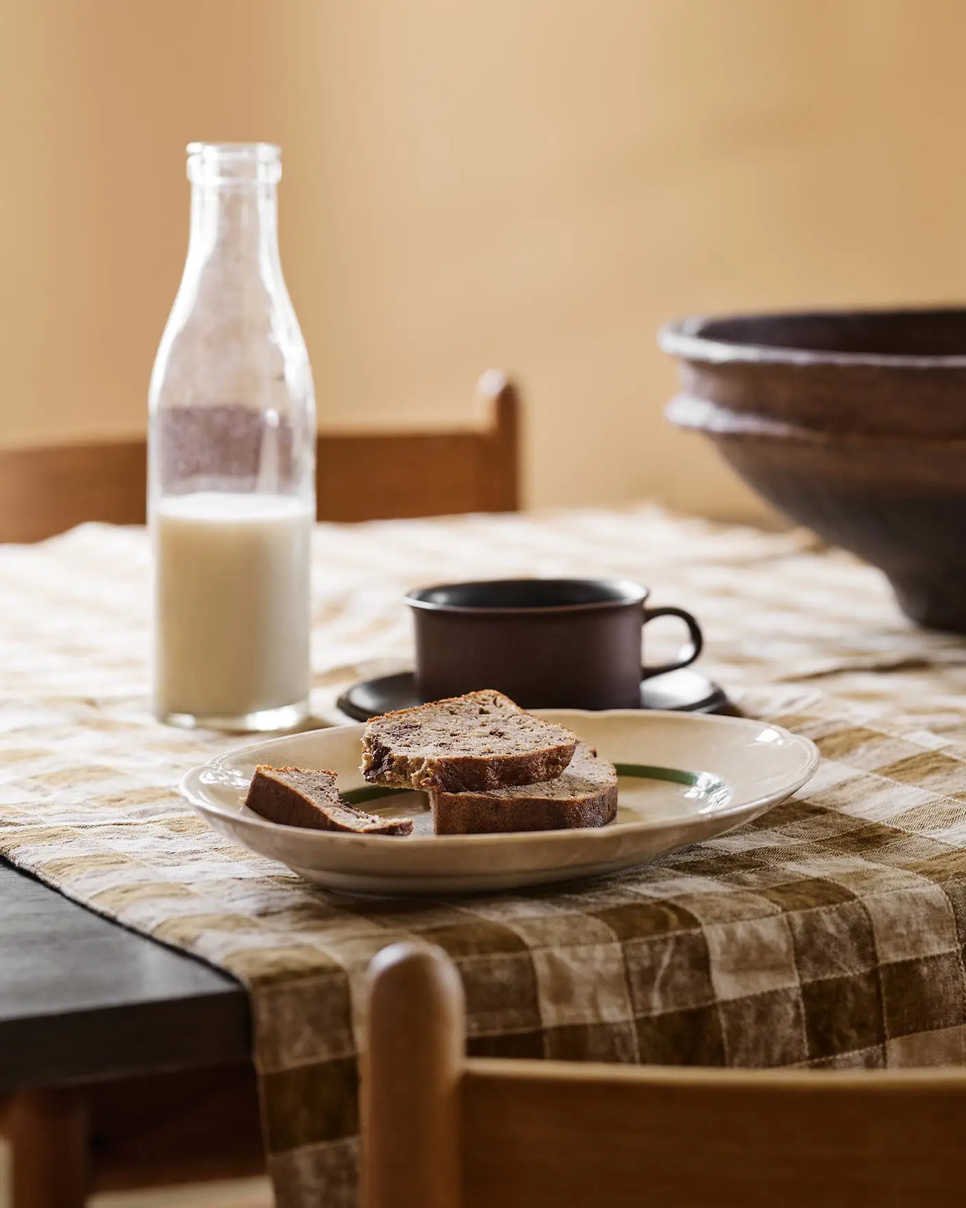 Table with bread, a cup, and a milk bottle on a checkered cloth against a ochre yellow background painted in with the Jotun colour 11202 Mild Ochre.
