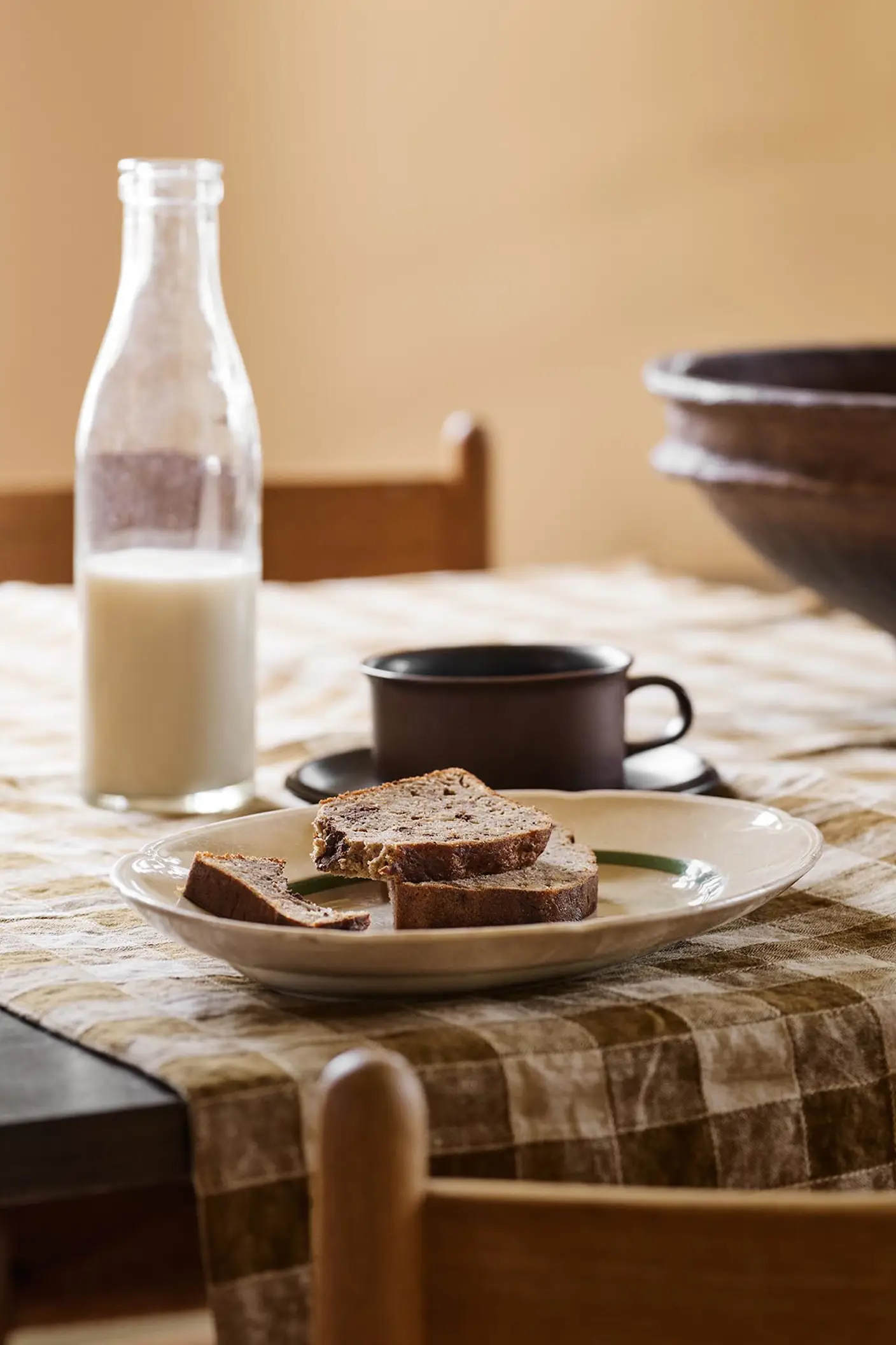 Table with bread, a cup, and a milk bottle on a checkered cloth against a ochre yellow background painted in with the Jotun colour 11202 Mild Ochre.