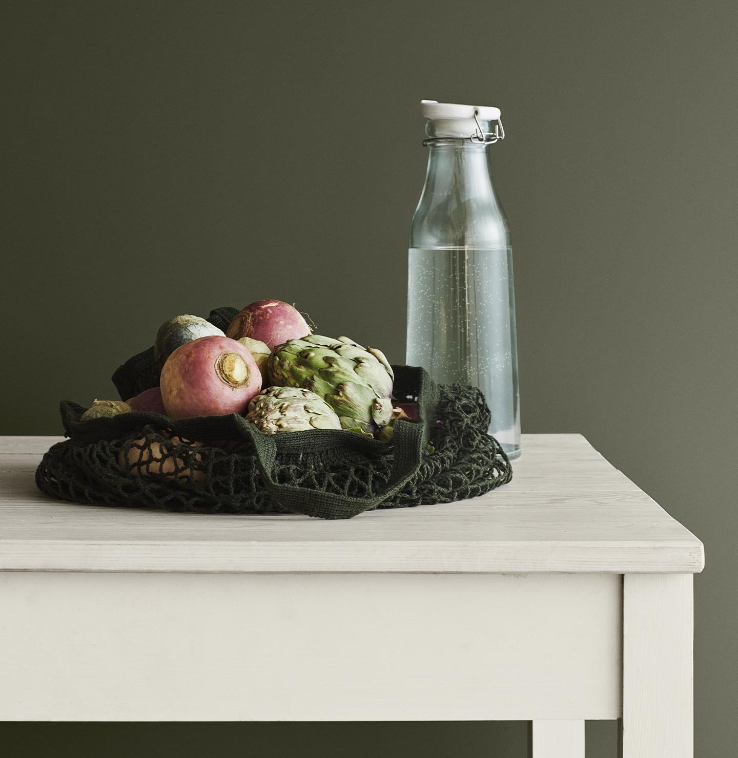 En nätpåse fylld med grönsaker på ett ljust träbord, mot en djup, jordnära grön vägg målad med Jotun 8494 Organic Green. Bordet är målat i färgen 1024 Tidlös från Jotun.