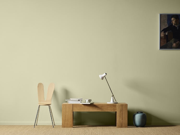 Bord i eik med en hvit lampe og en moderne stol ved siden av, alt mot en lys grønn vegg malt i Jotun Sunny Green