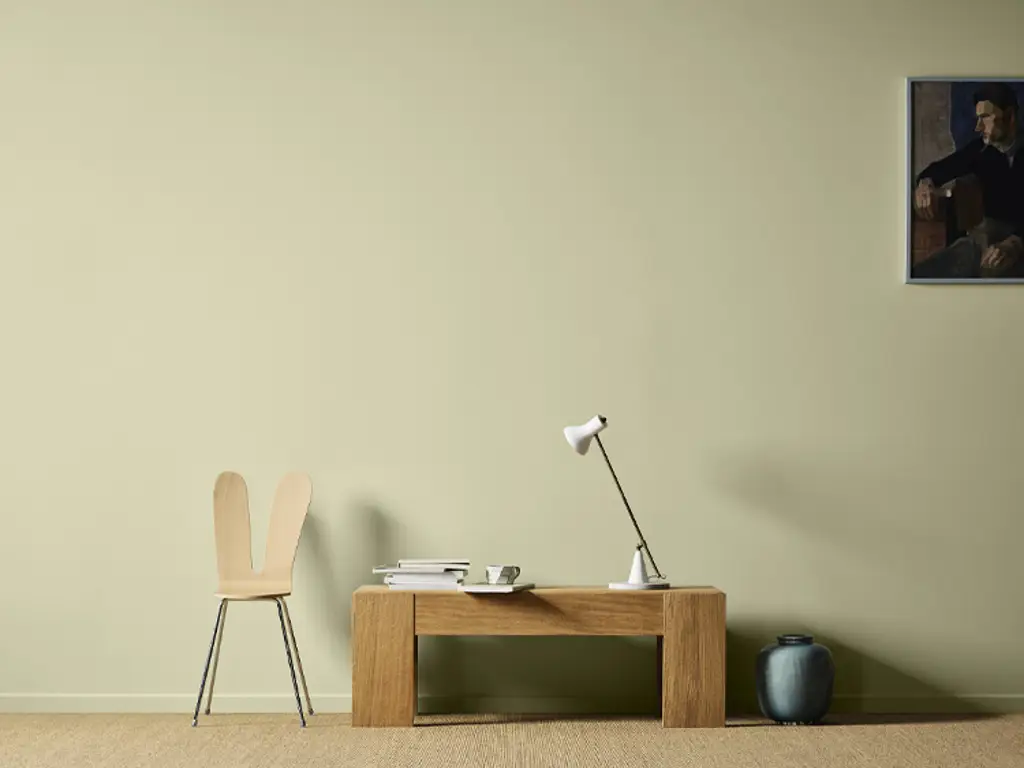 Bord i eik med en hvit lampe og en moderne stol ved siden av, alt mot en lys grønn vegg malt i Jotun Sunny Green