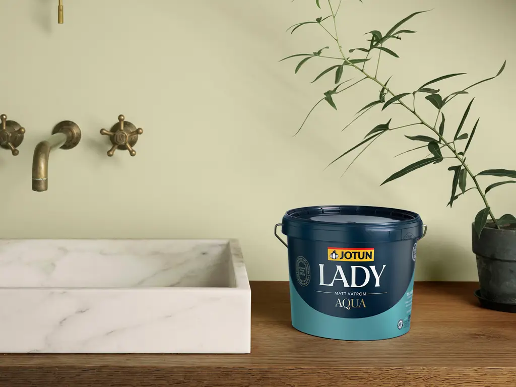 JOTUN LADY Aqua malingbøtte ved en hvit marmorkum på et trebord, med plante og messingkran i bakgrunnen