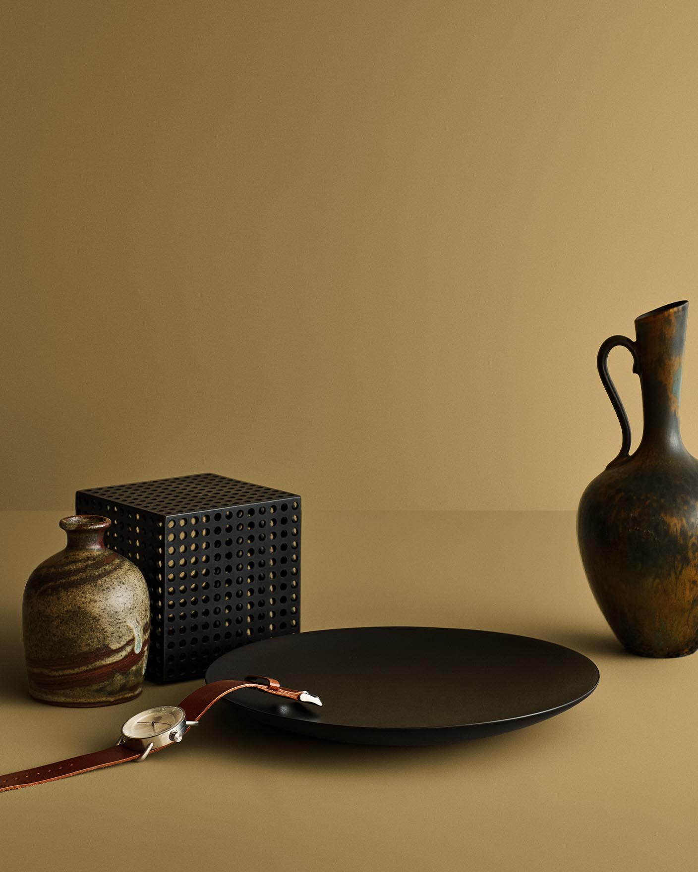 Stilleben med keramisk vase, svart tallerken og armbåndsur mot en beige gul bakgrunn malt med Jotuns farge 11174 Curious Mind.
