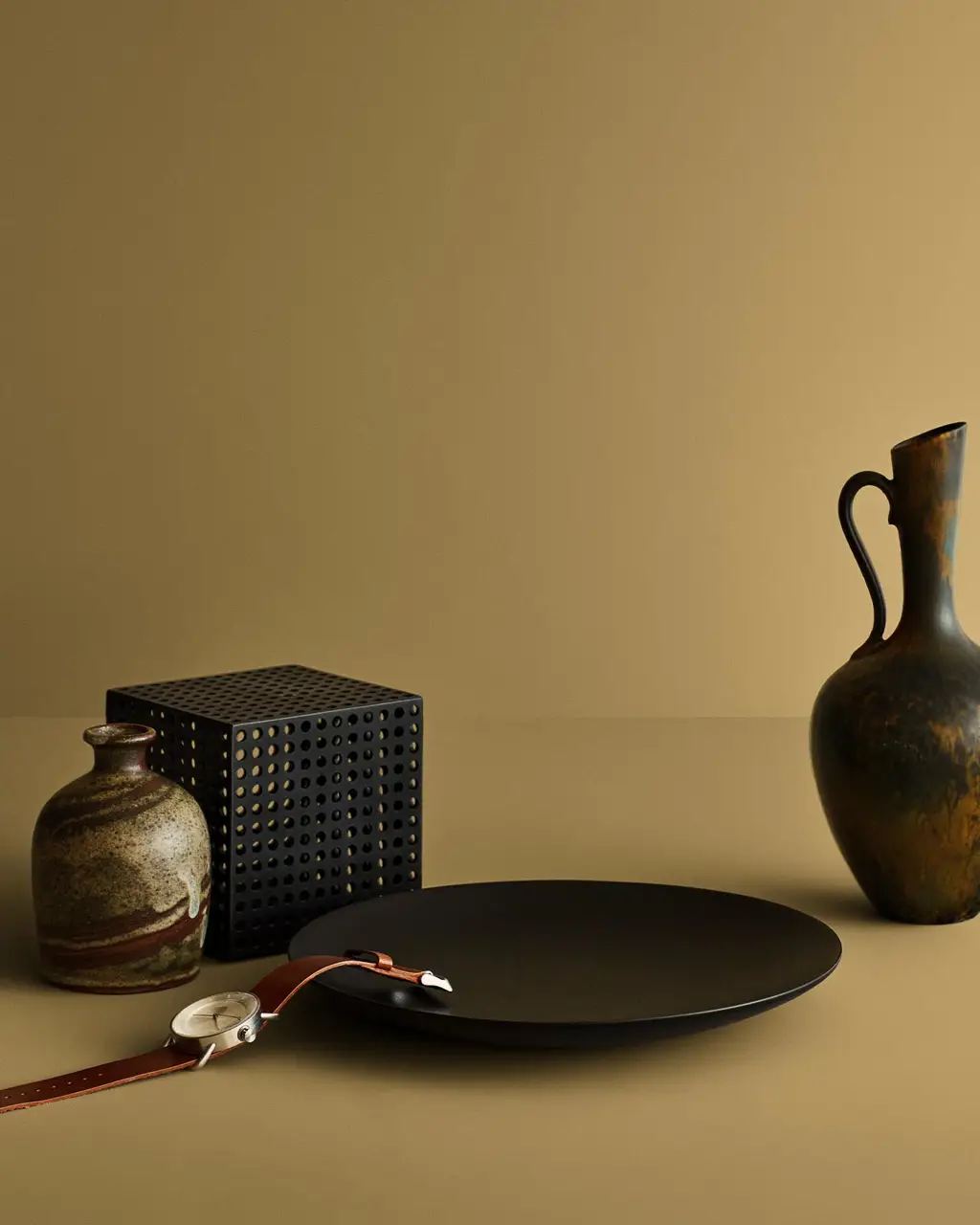 Stilleben med keramisk vase, svart tallerken og armbåndsur mot en beige gul bakgrunn malt med Jotuns farge 11174 Curious Mind.