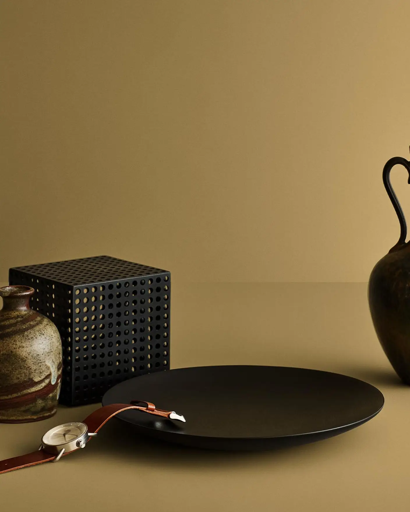 Still life with ceramic vase, black plate, and wristwatch against a beige yellow background painted by Jotun's colour 11174 Curious Mind.