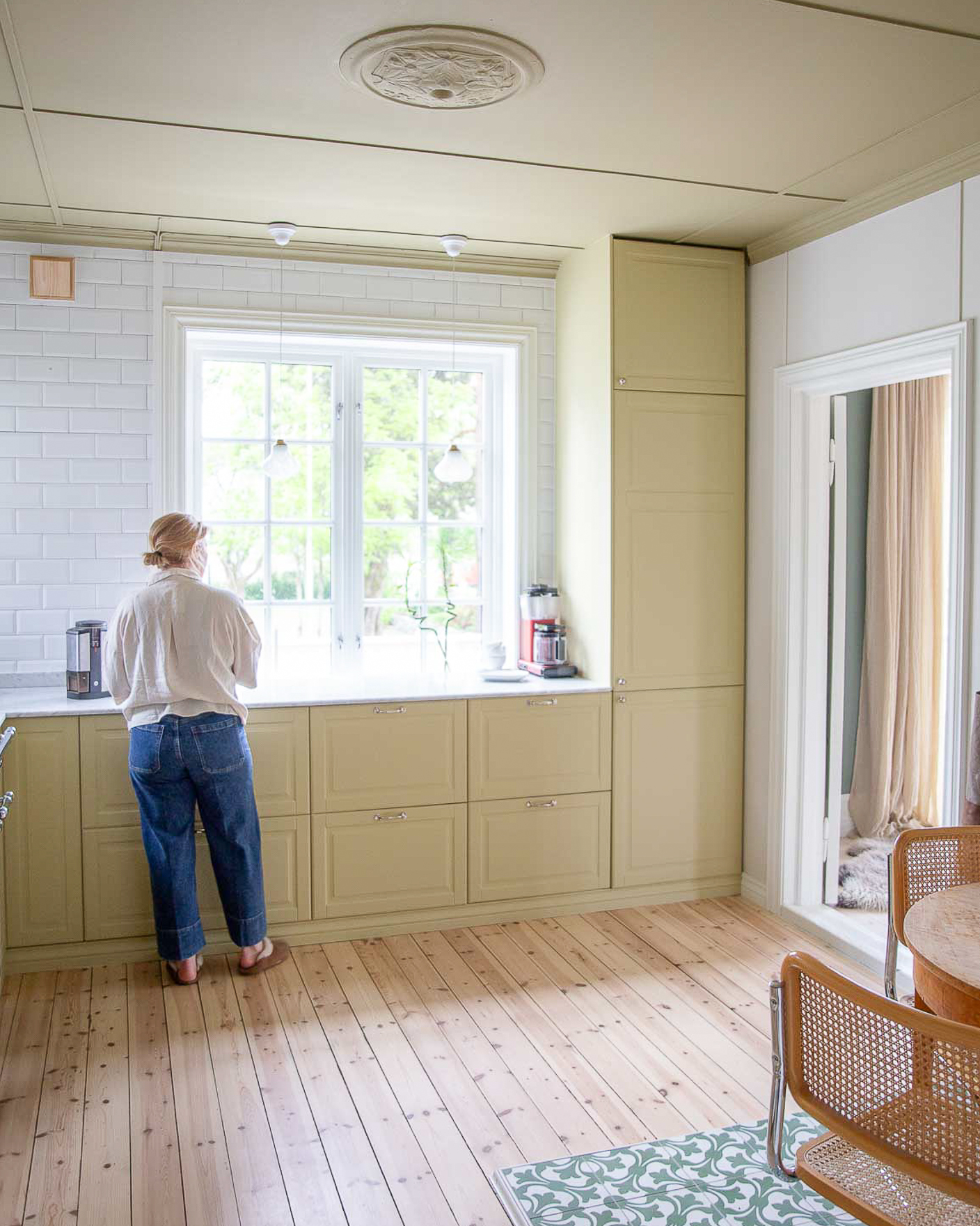 Kvinne ved kjøkkenbenk på gulmalt kjøkken i Jotun Earthy Yellow, tak malt mørkere gult med Jotun Khaki Yellow, vegg i gråhvite Jotun Frostrøyk.