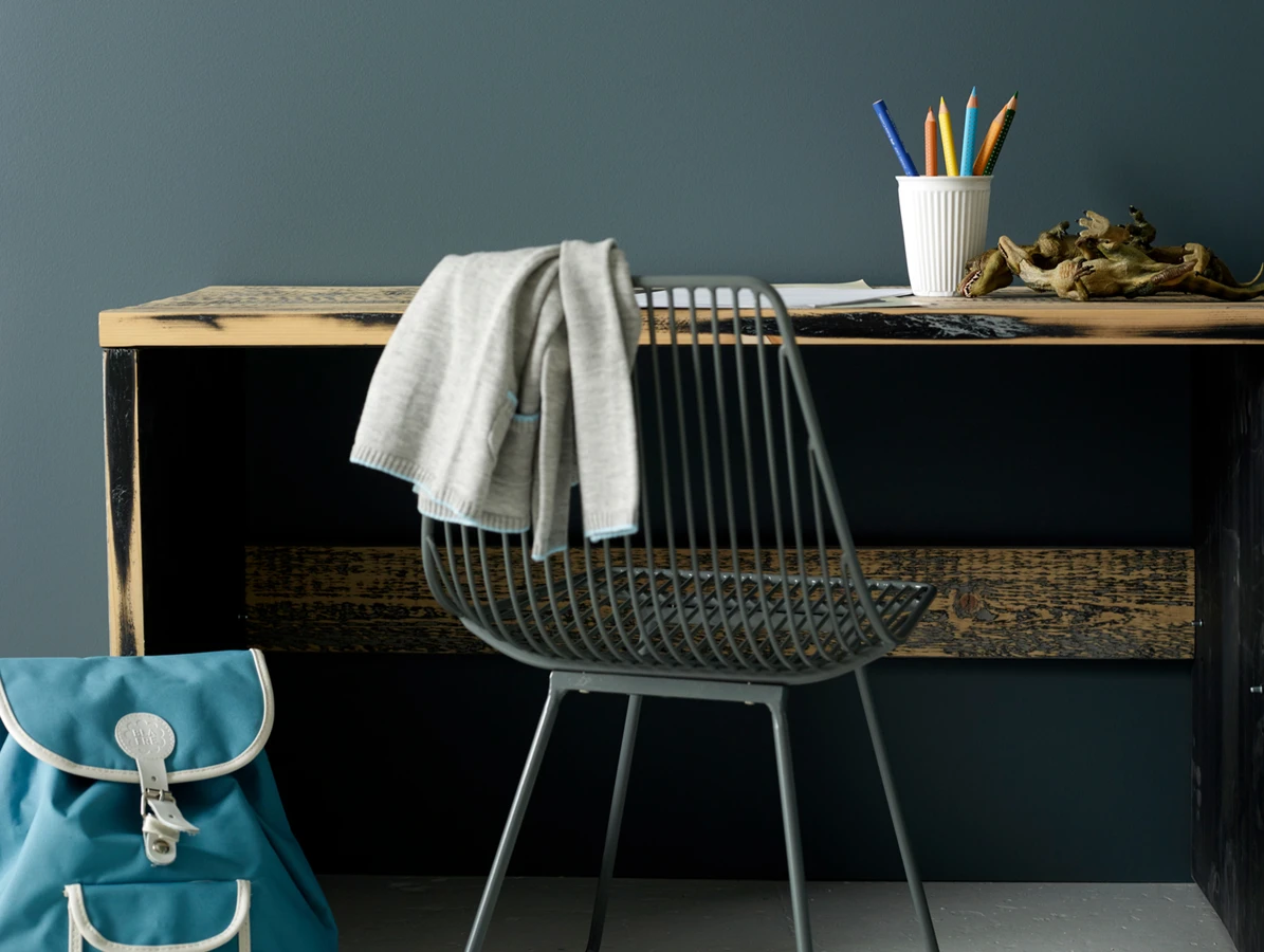Modern desk with wire chair, draped sweater, pencils in cup, and blue backpack in a child playroom.