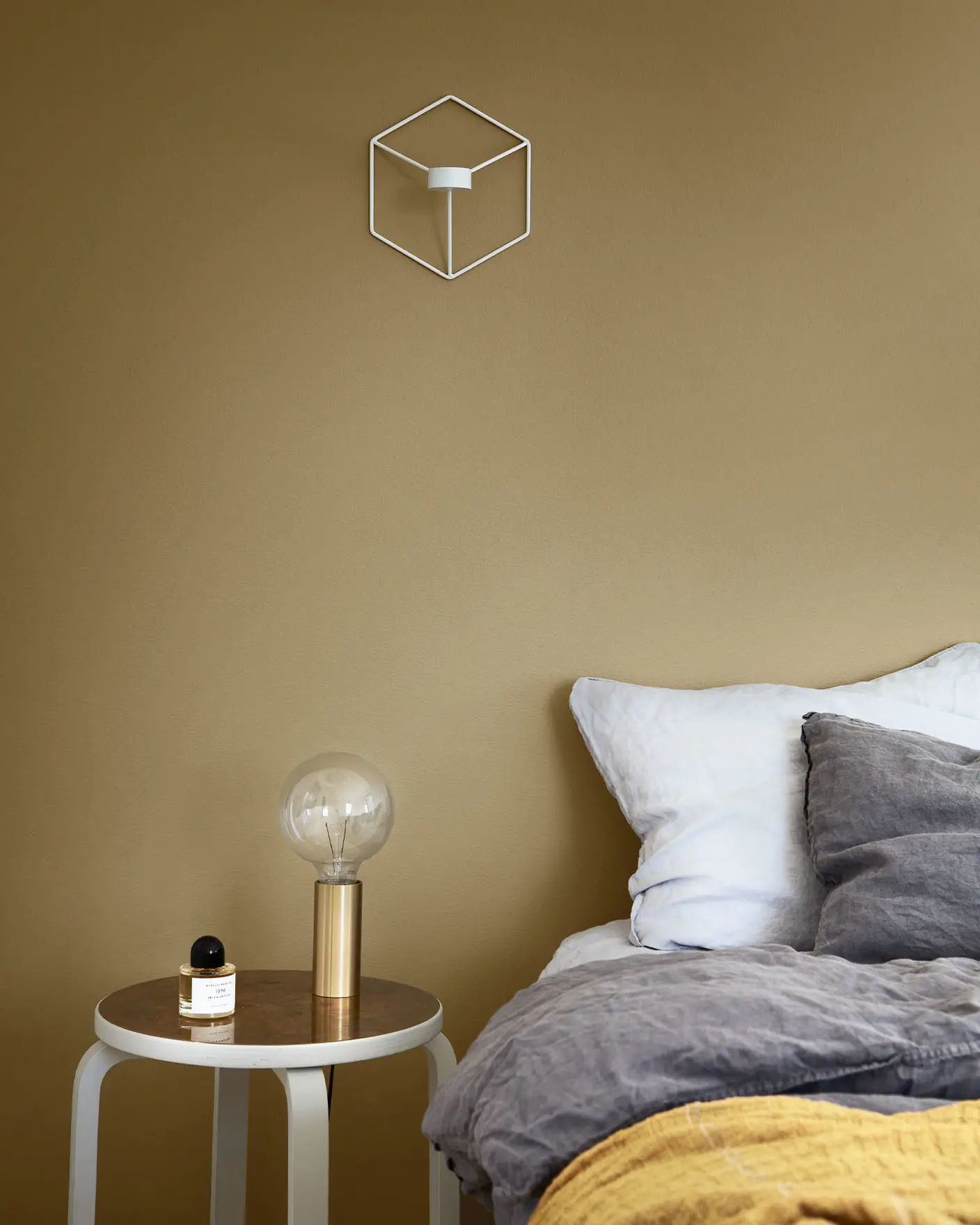 Mysig sovrumshörna med ett grått och senapsgult bäddset, ett litet runt sidobord med en guldlampa och en parfymflaska, mot en dämpad gulgrön vägg målad med Jotun 1864 Anonymous.