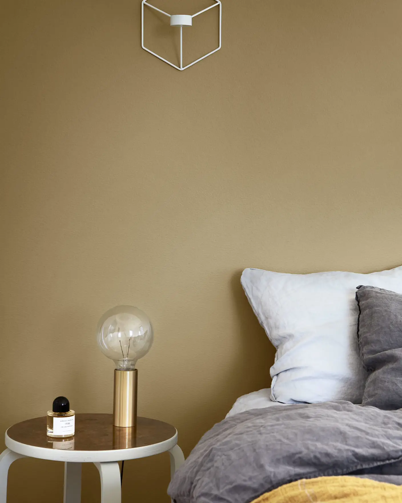 Mysig sovrumshörna med ett grått och senapsgult bäddset, ett litet runt sidobord med en guldlampa och en parfymflaska, mot en dämpad gulgrön vägg målad med Jotun 1864 Anonymous.