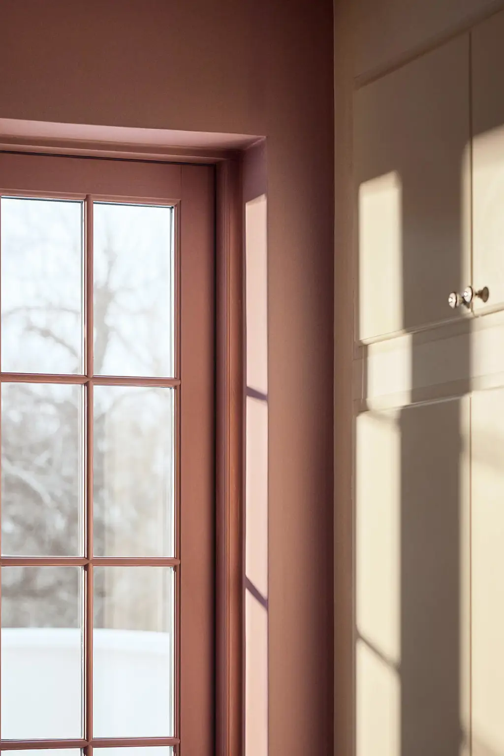 En stue med et vindue malet i den rosa farve Jotun 20256 Balmy Pink og tilstødende skabe i den gyldenbeige Jotun 11365 Morning Glow, oplyst af blødt sollys.