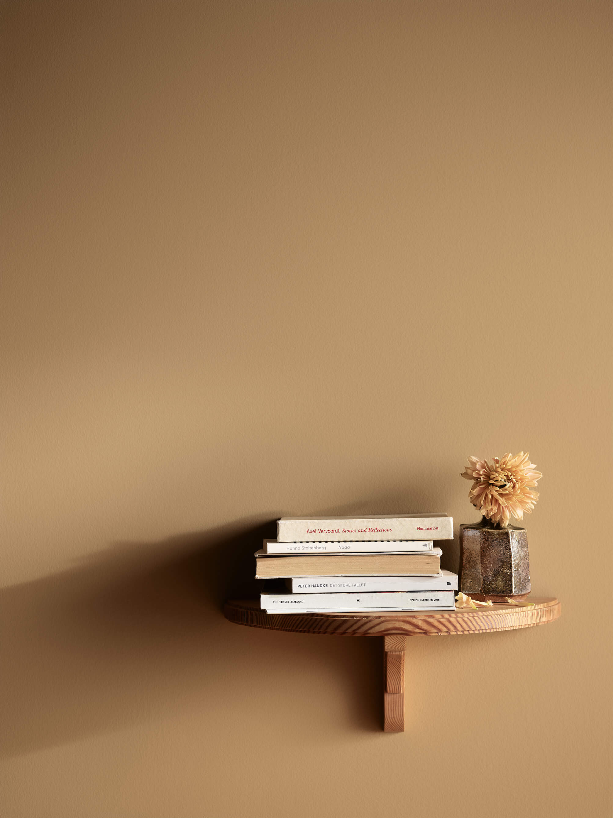 En stue med en vegg malt okergul i Jotun 1909 Chalk Ochre, med en liten vegghylle i tre med en bokstabel og en vase med en blomst