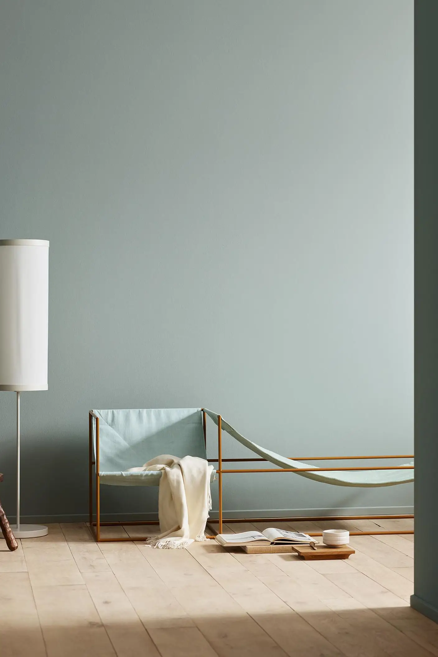 Minimalist bedroom with a light blue chair, white blanket, and wooden stool against a muted blue wall painted with the colour 6378 Iconic from Jotun.