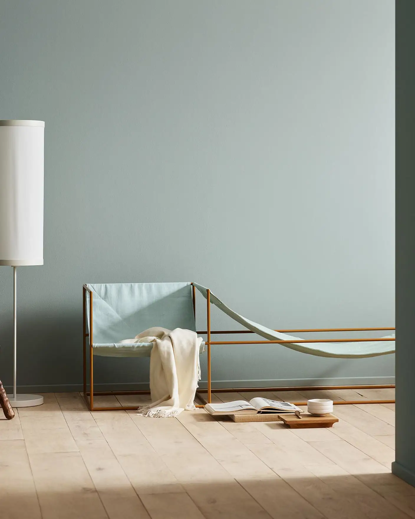 Minimalist bedroom with a light blue chair, white blanket, and wooden stool against a muted blue wall painted with the colour 6378 Iconic from Jotun.