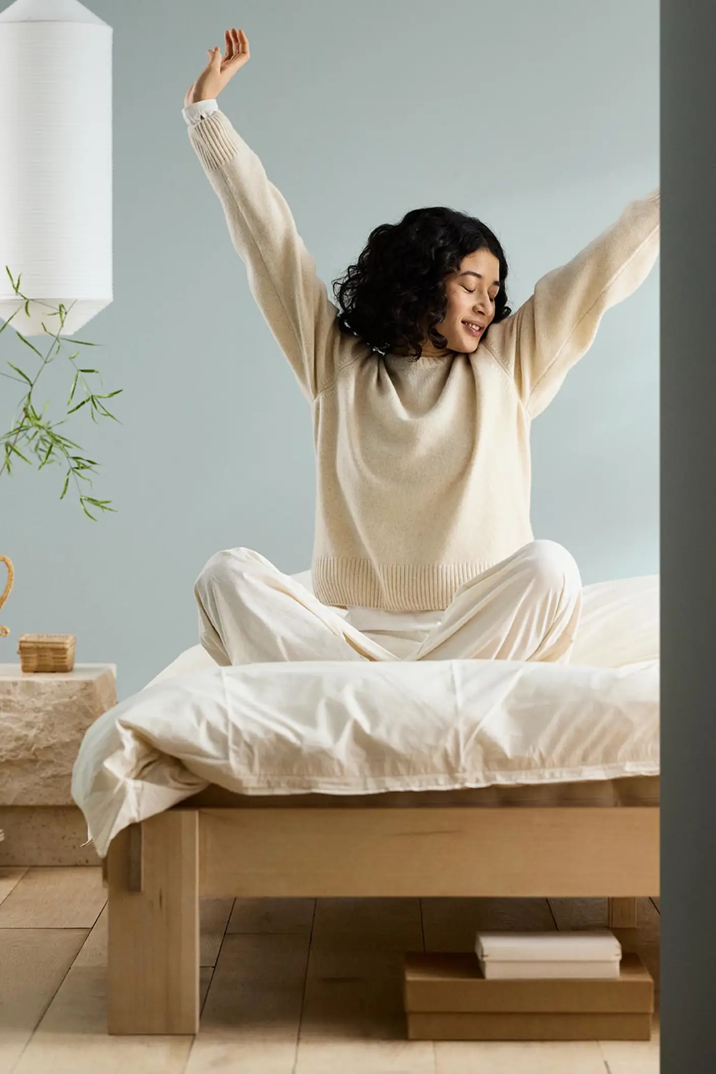 Woman sitting on a bed with arms raised, next to a bedside table against a muted blue wall painted in the Jotun colour 6378 Iconic.