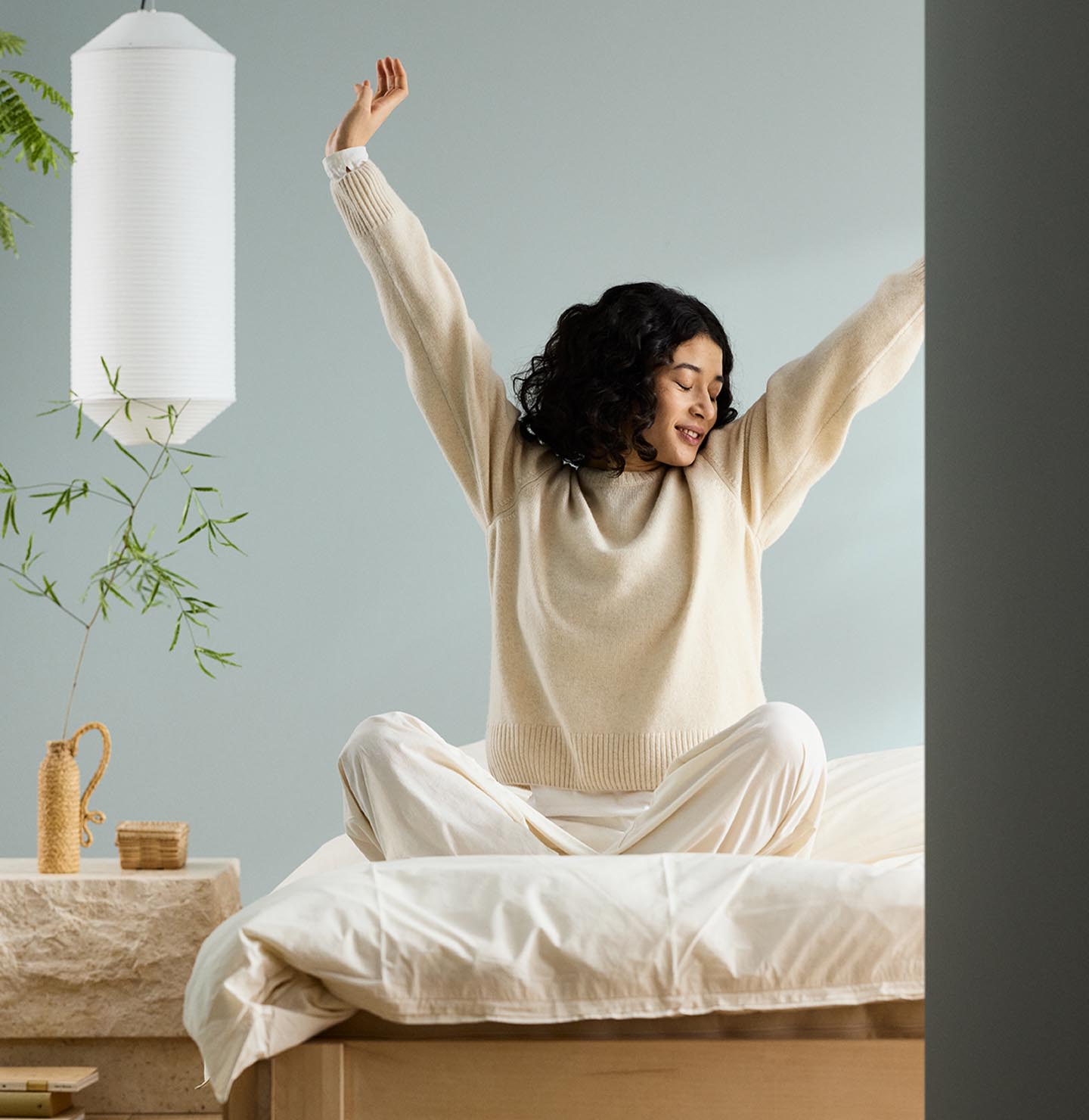 Woman sitting on a bed with arms raised, next to a bedside table against a muted blue wall painted in the Jotun colour 6378 Iconic.