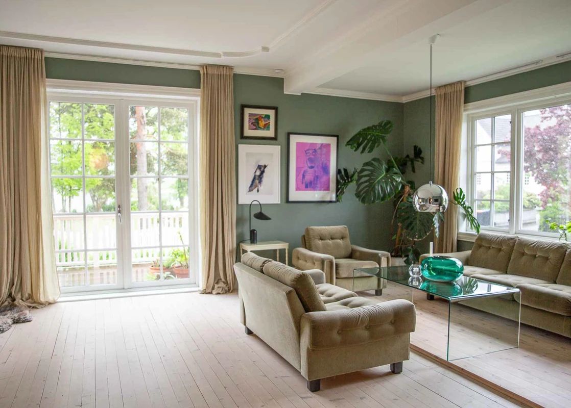 Stue med stor terrassedør og vindu med sprosser, lyst tregulb og vegger malt grønne i Jotun LADY Antique Green, tak med ornamenter malt hvitt i fargen Bomull fra Jotun. Grønnbeige salong, glassbord og beige gardiner.