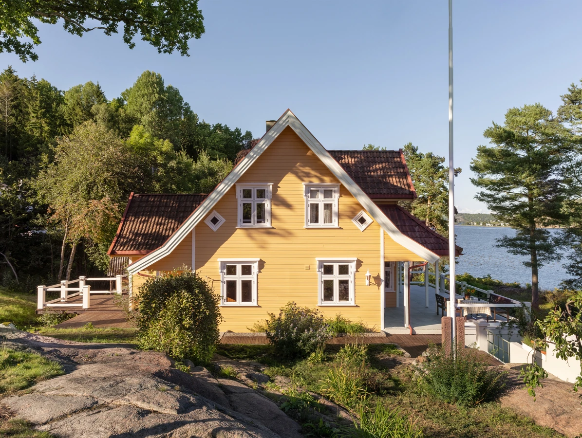 Gult trehus malt i fargen Skagenul fra Jotun ved vannet omgitt av skog og svaberg, med hvite vinduer og rød takstein.