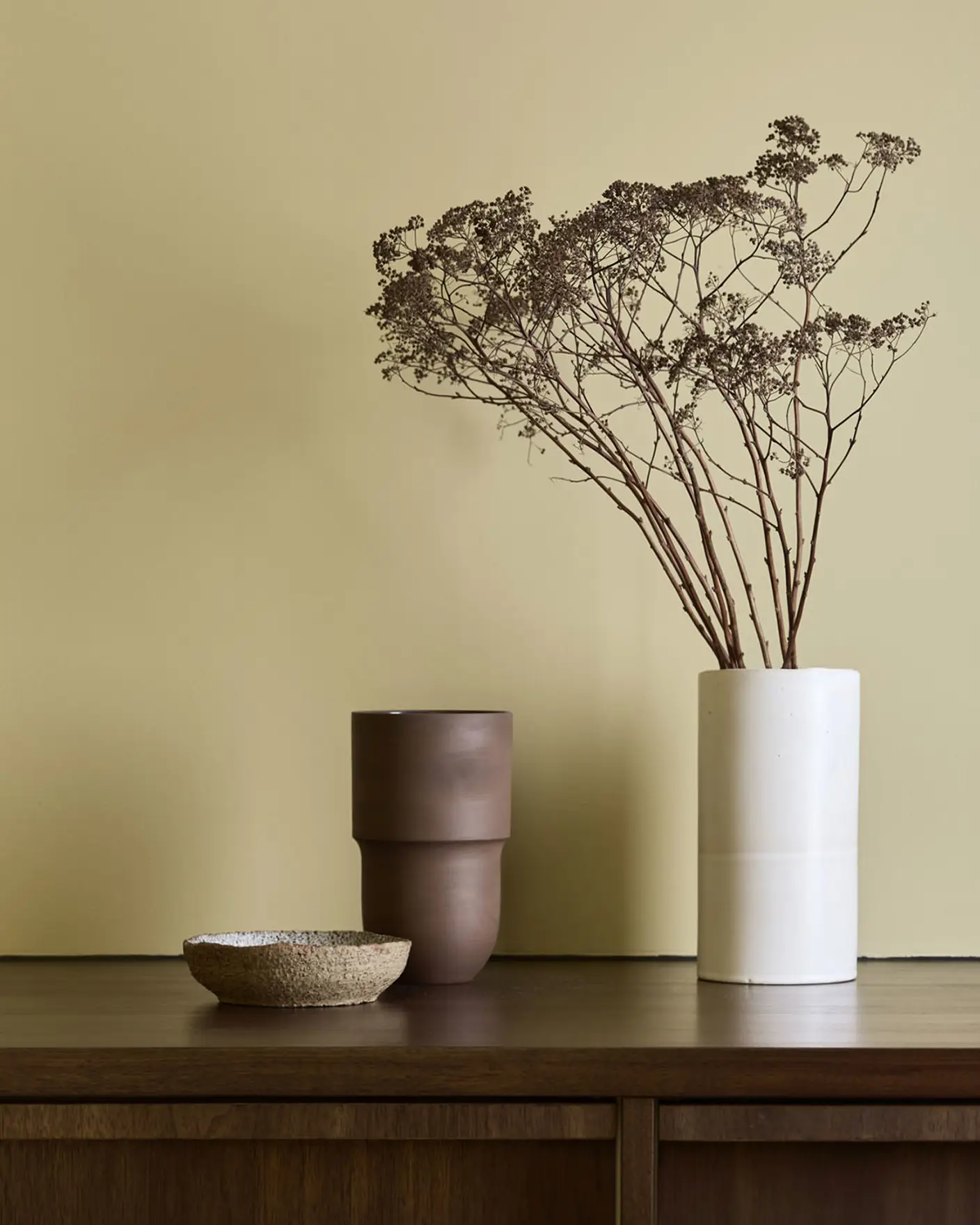 Keramikk og en blomstervase som står på toppen av et kjøkkenskap i mørkt valnøttre. Veggene er malt i Jotun Earthy Yellow