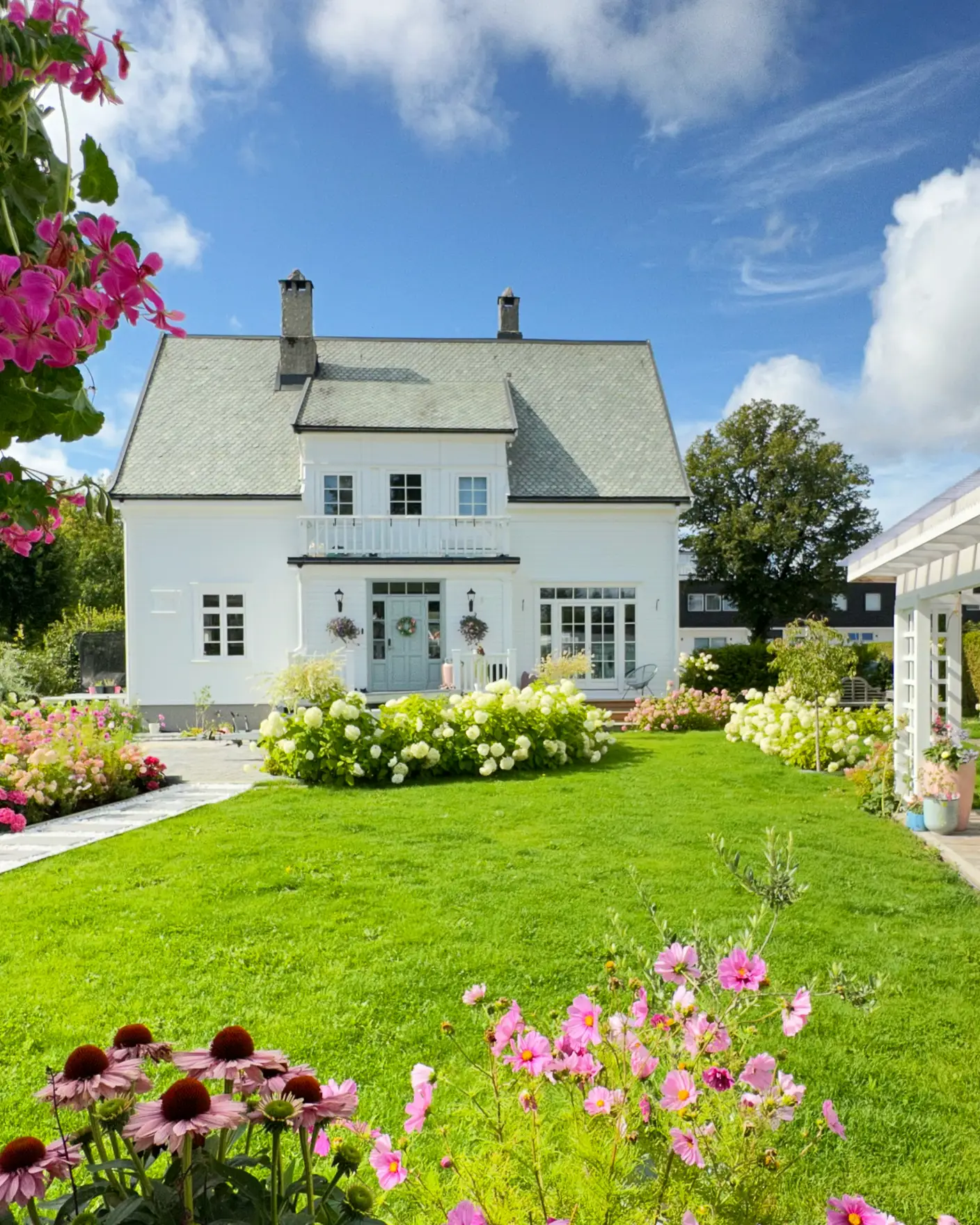 Hvitt trehus malt i Jotun 9918 Klassisk Hvit med DRYGOLIN Power Clean, grønn og blomstrende hage i front, grønt gress og bed.