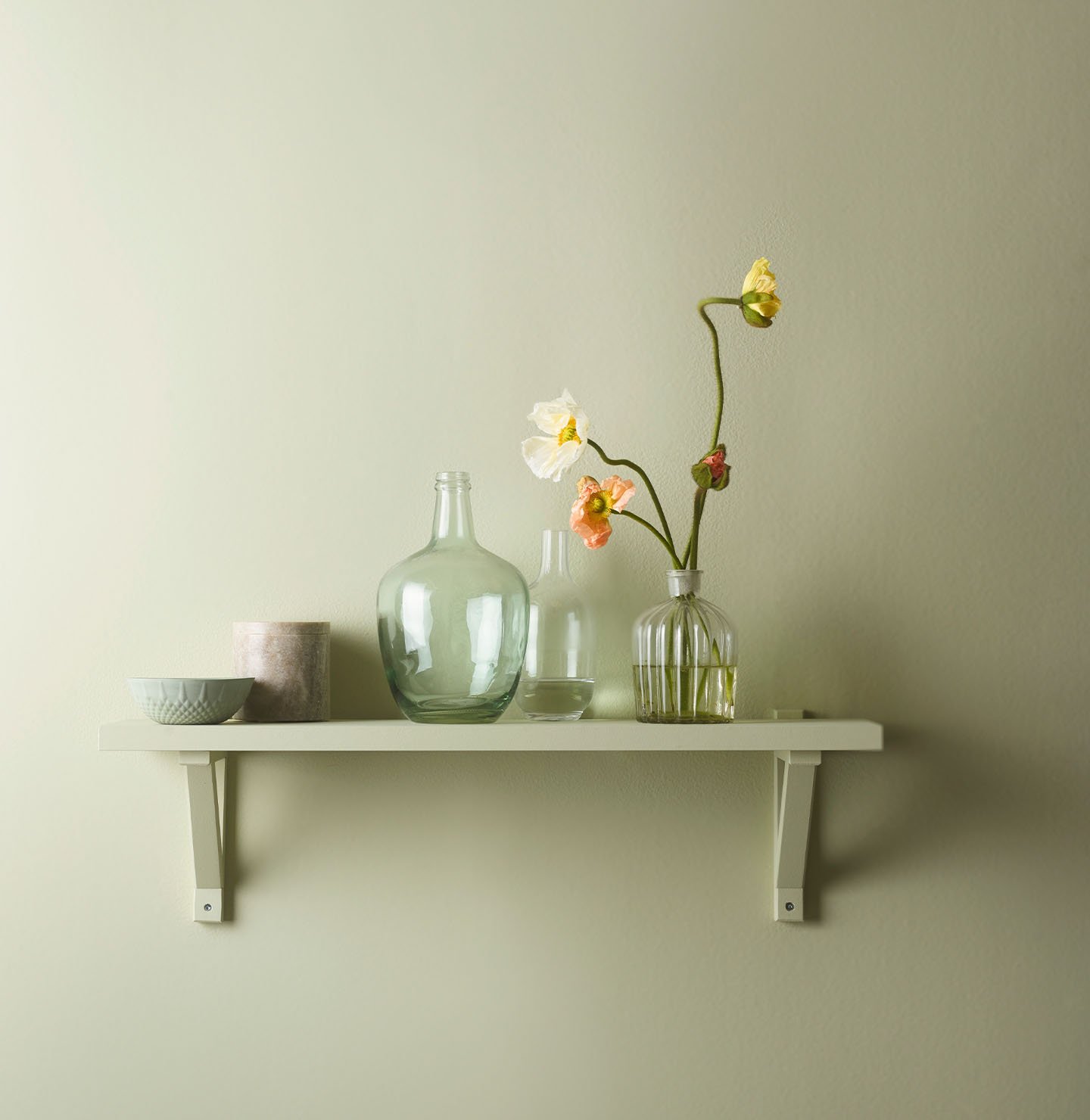 Hylla på väggen inredd med vackra glasvaser och blommor, mot en gulgrön vägg målad med Jotun 8222 Kryddgrönt för ett fräscht och lätt uttryck.