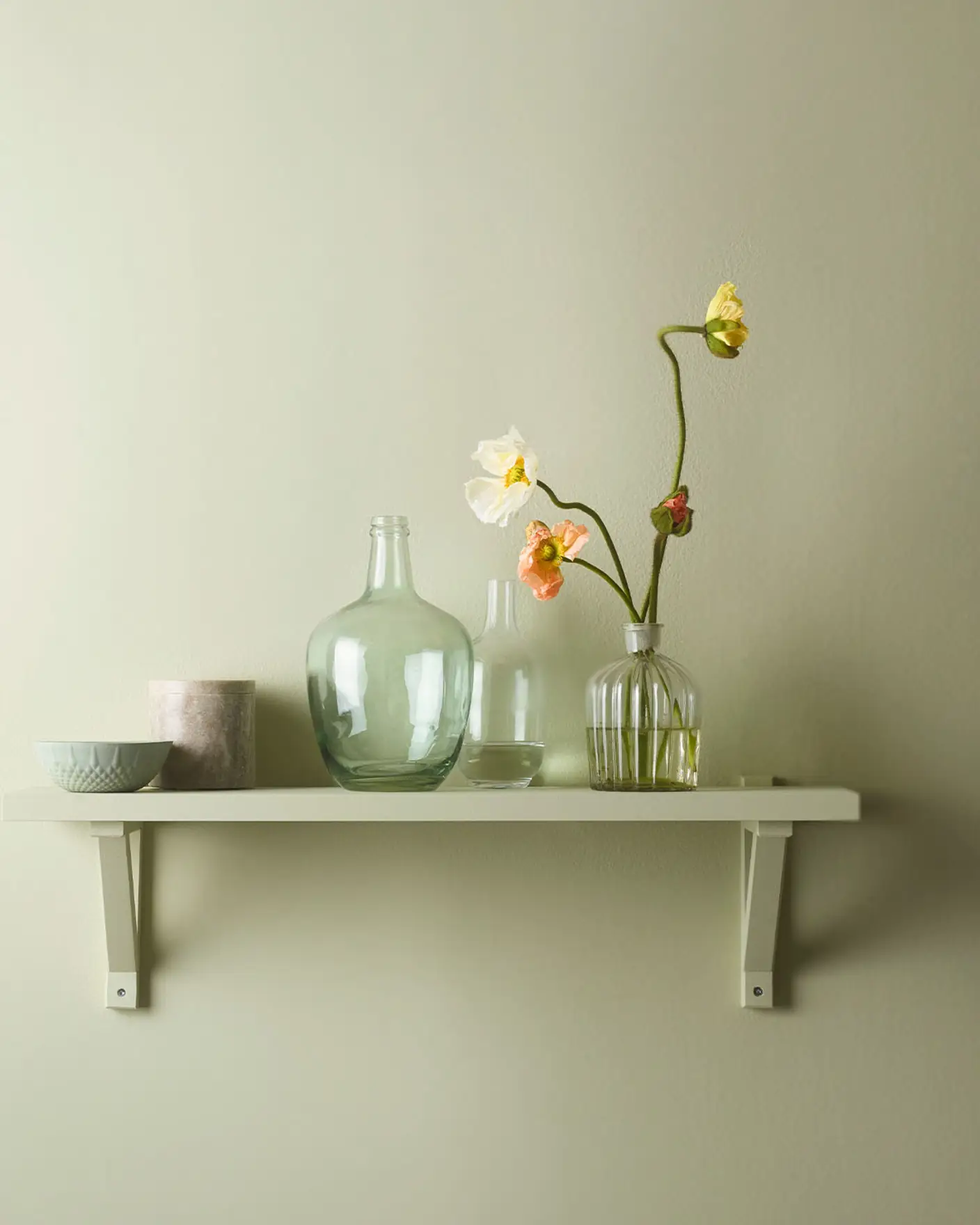 Hylla på väggen inredd med vackra glasvaser och blommor, mot en gulgrön vägg målad med Jotun 8222 Kryddgrönt för ett fräscht och lätt uttryck.