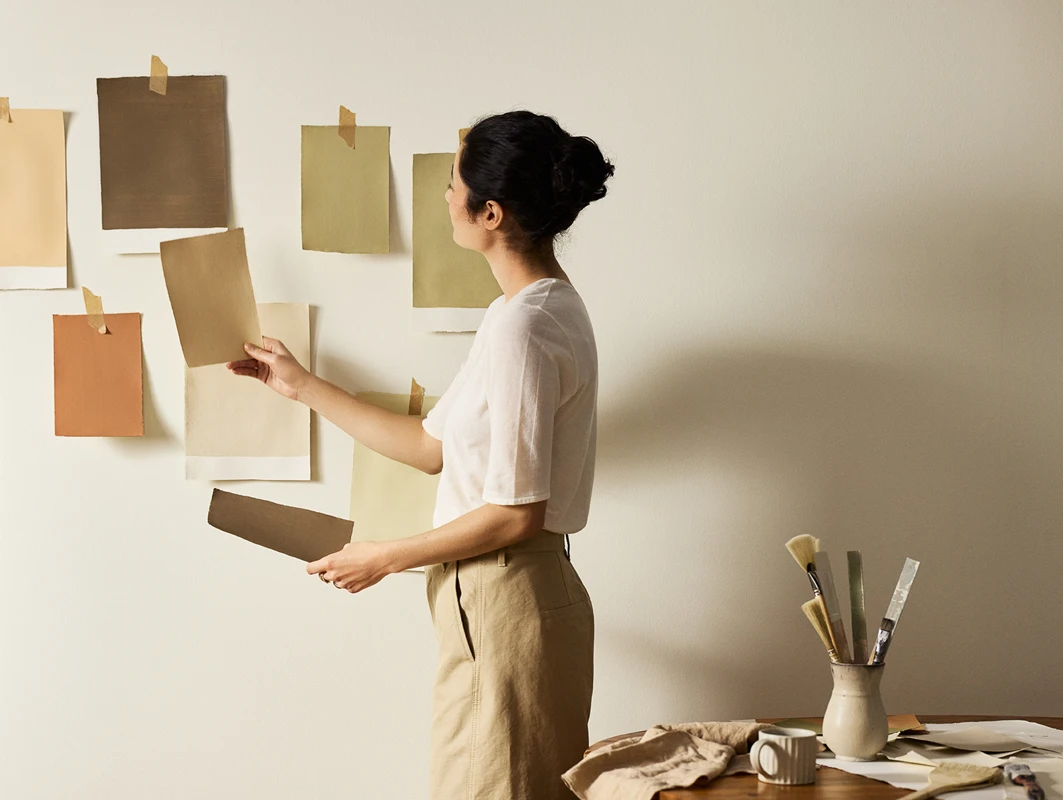 Kvinne henger opp fargeprøver malt med Jotun-farger på en beige vegg malt i Jotun Soul