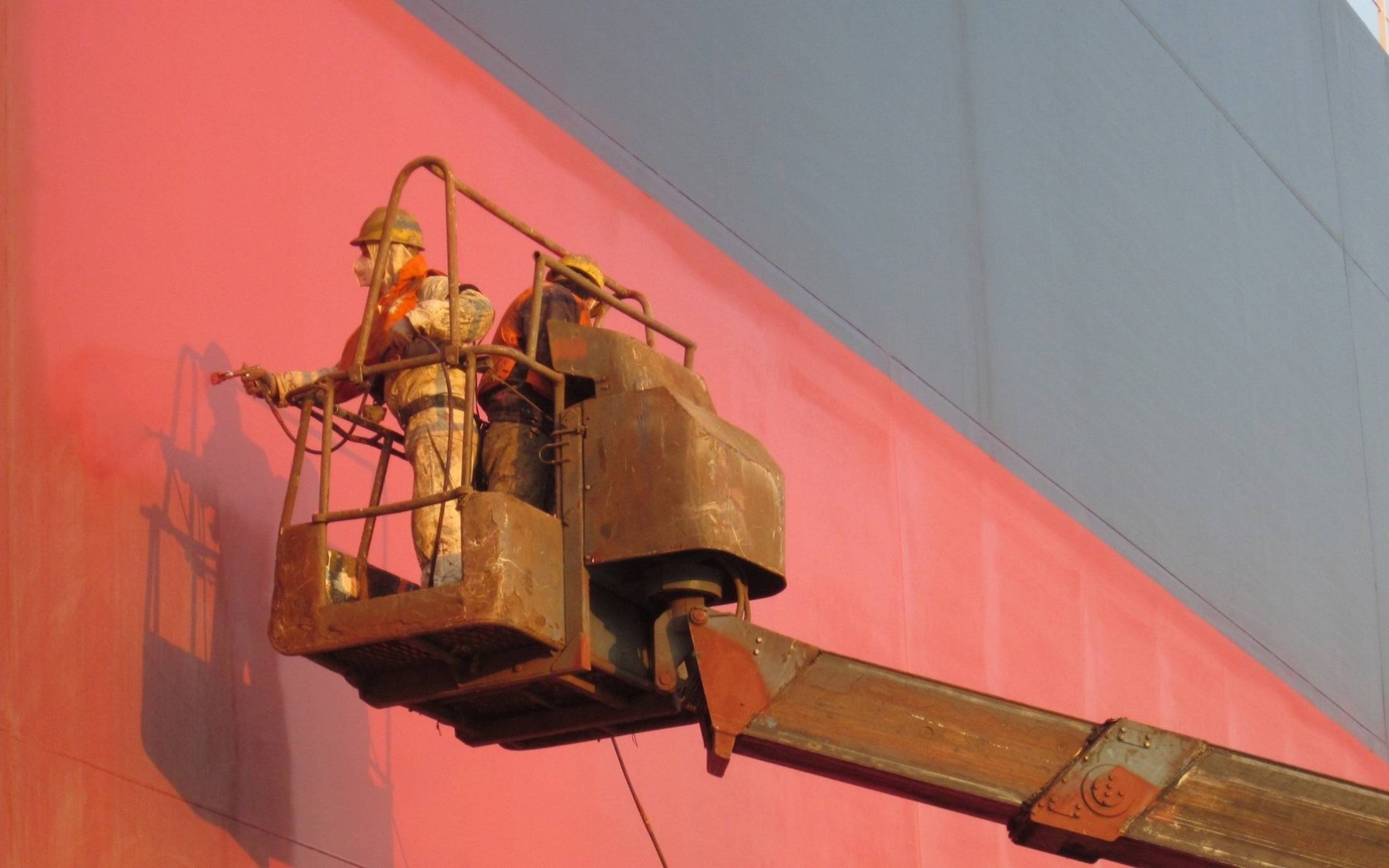 Two applicators spray painting red ship hull