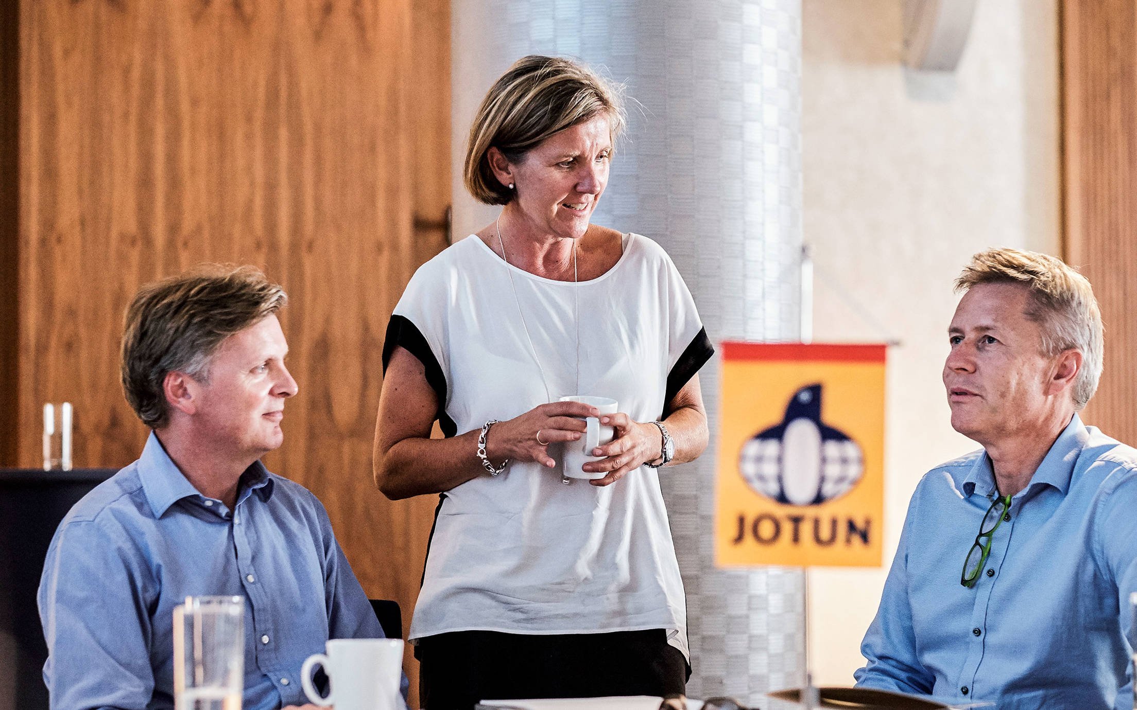 Two men with blue shirts sitting and a woman standing holding a coffee cup