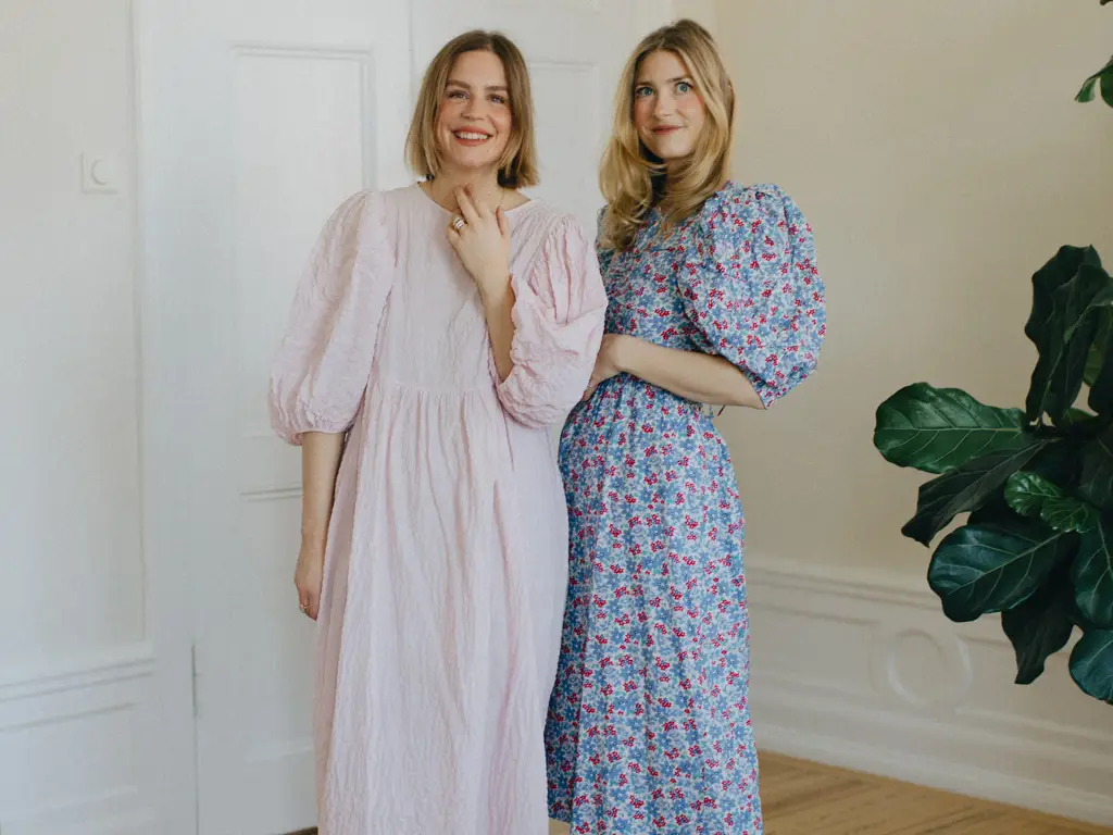Sofia Wood och Elsa Billgren i sommarklänningar står i ett ljust rum, en i rosa klänning och en i blommig klänning, bredvid en stor krukväxt