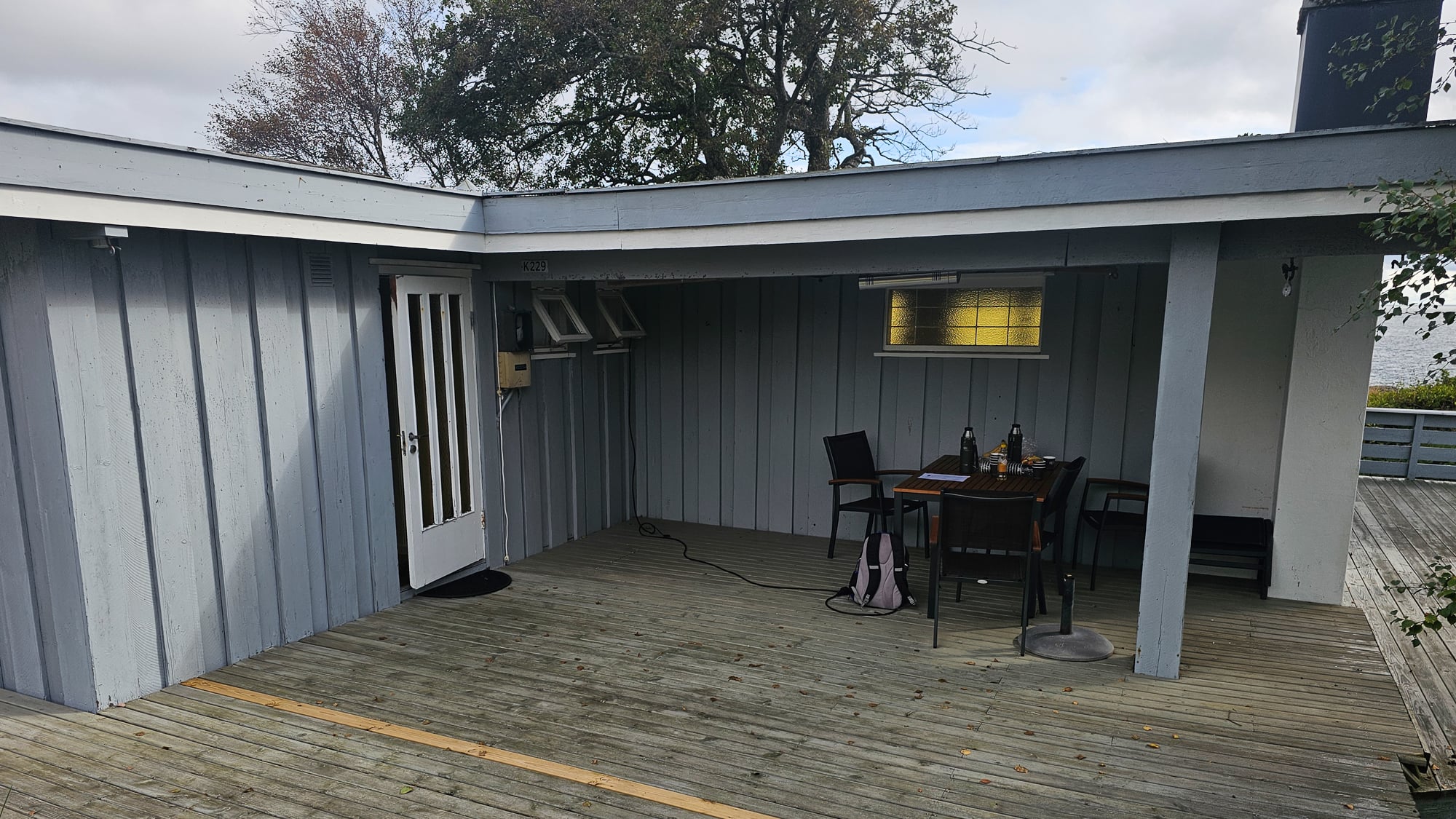 Hytteterrasse med overbygg før, grå terrasse og slitne blå vegger med et lite bord.