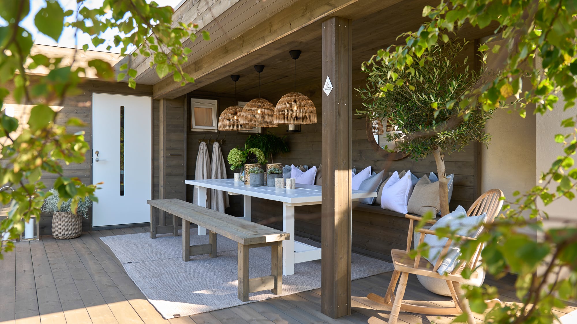 Hytteterrasse etter: med nytt terrassegulv, overbygg, benk med puter og stort bord med lamper over.