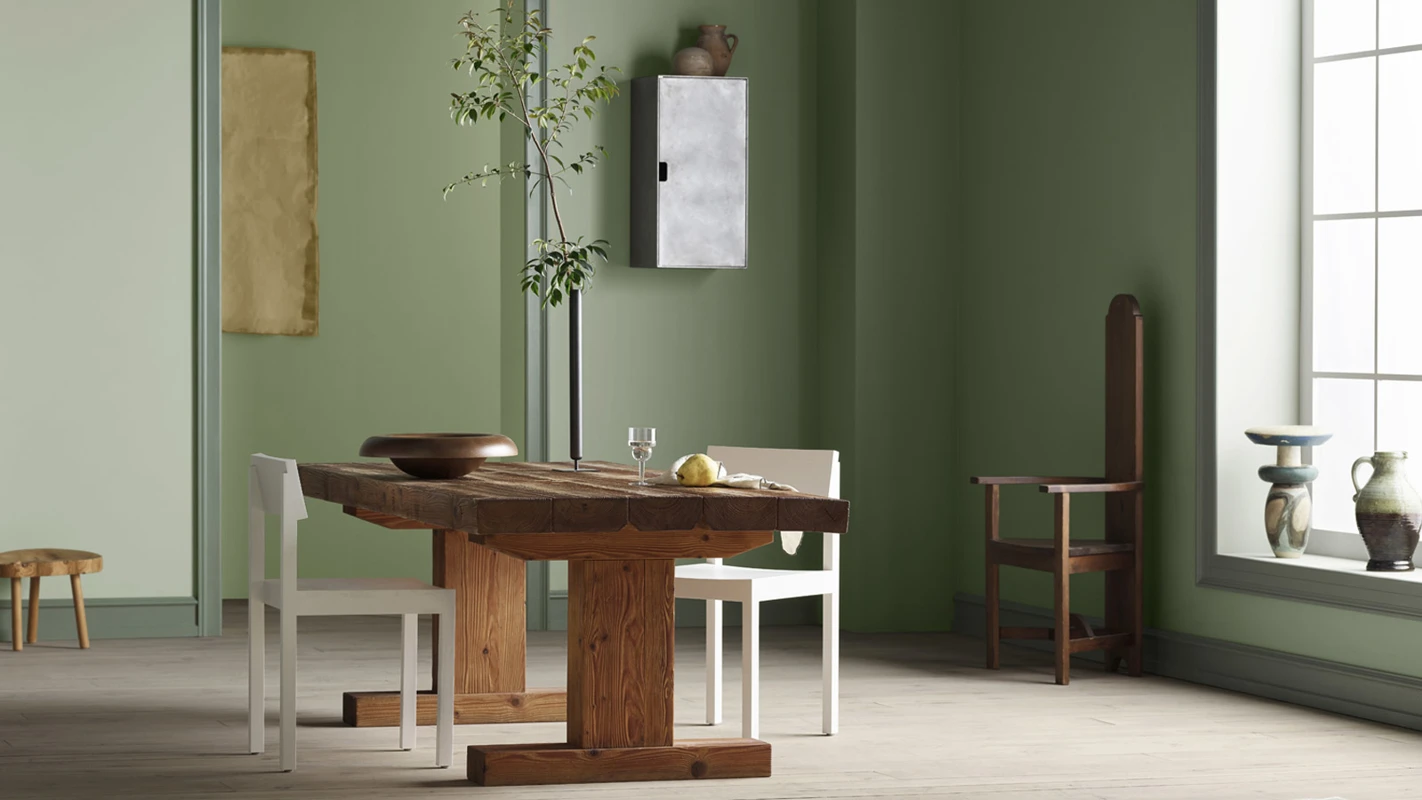 A minimalist room with sage green walls, a rustic wooden dining table with white chairs, and a window on the right