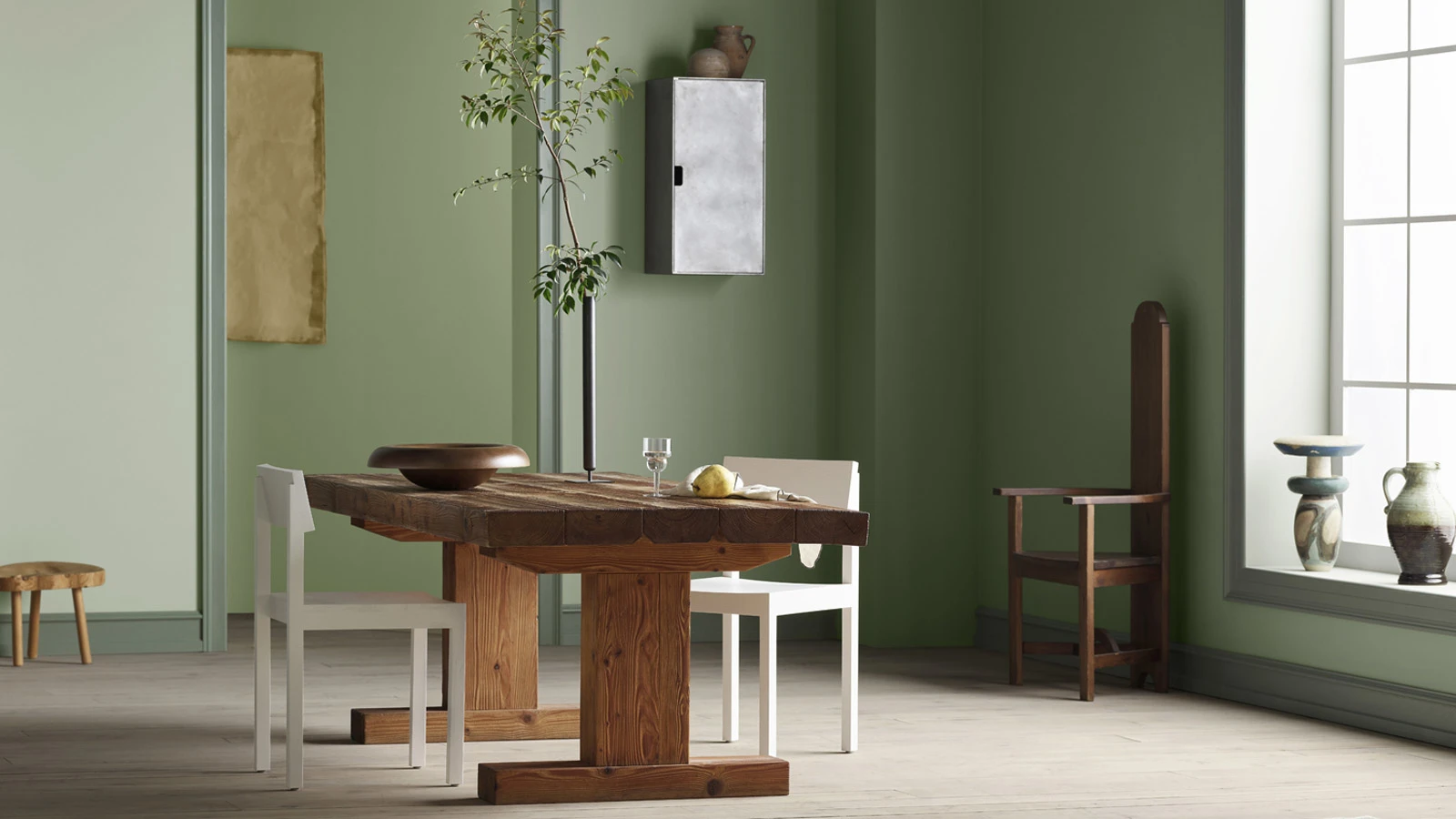 A minimalist room with sage green walls, a rustic wooden dining table with white chairs, and a window on the right