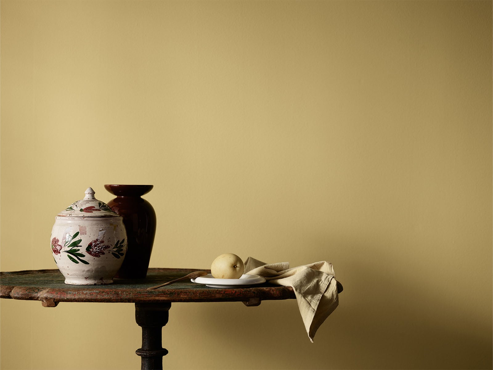 Stillben av keramikkrukker, tallerken med pære og beige duk på et rustikt bord mot en gul vegg malt i fargen Jotun Retro.