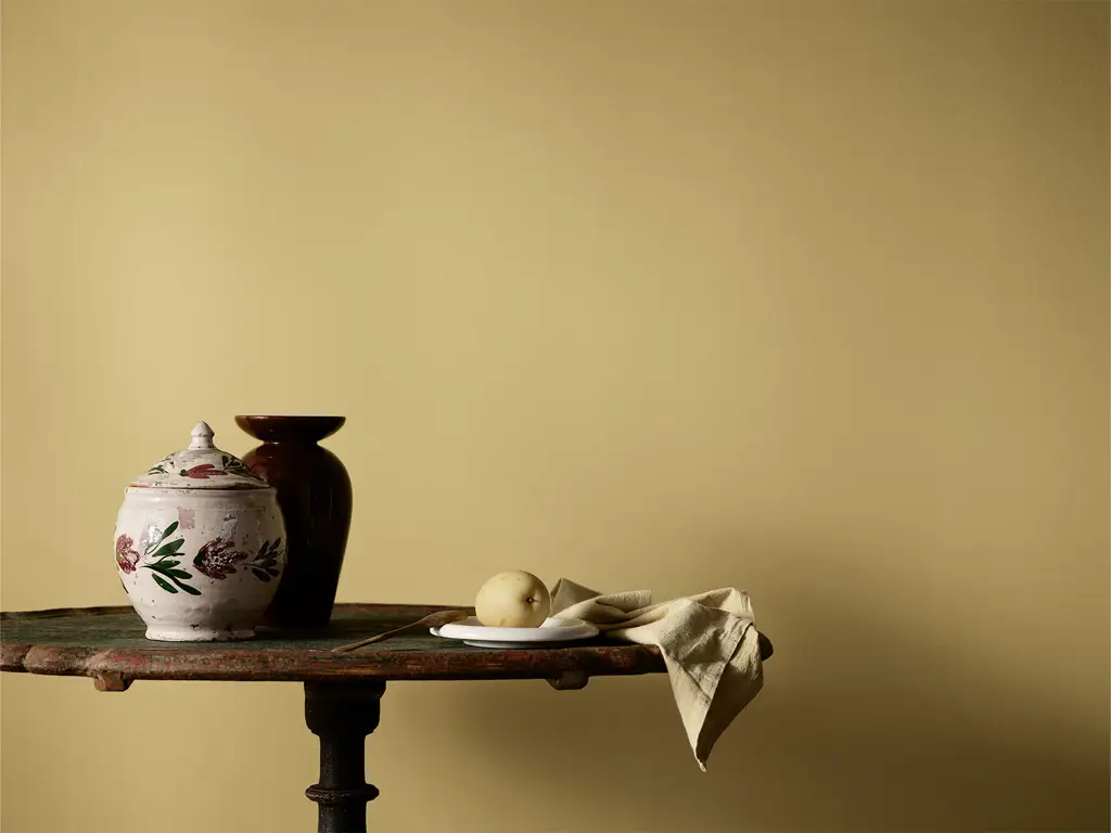 Stillben av keramikkrukker, tallerken med pære og beige duk på et rustikt bord mot en gul vegg malt i fargen Jotun Retro.