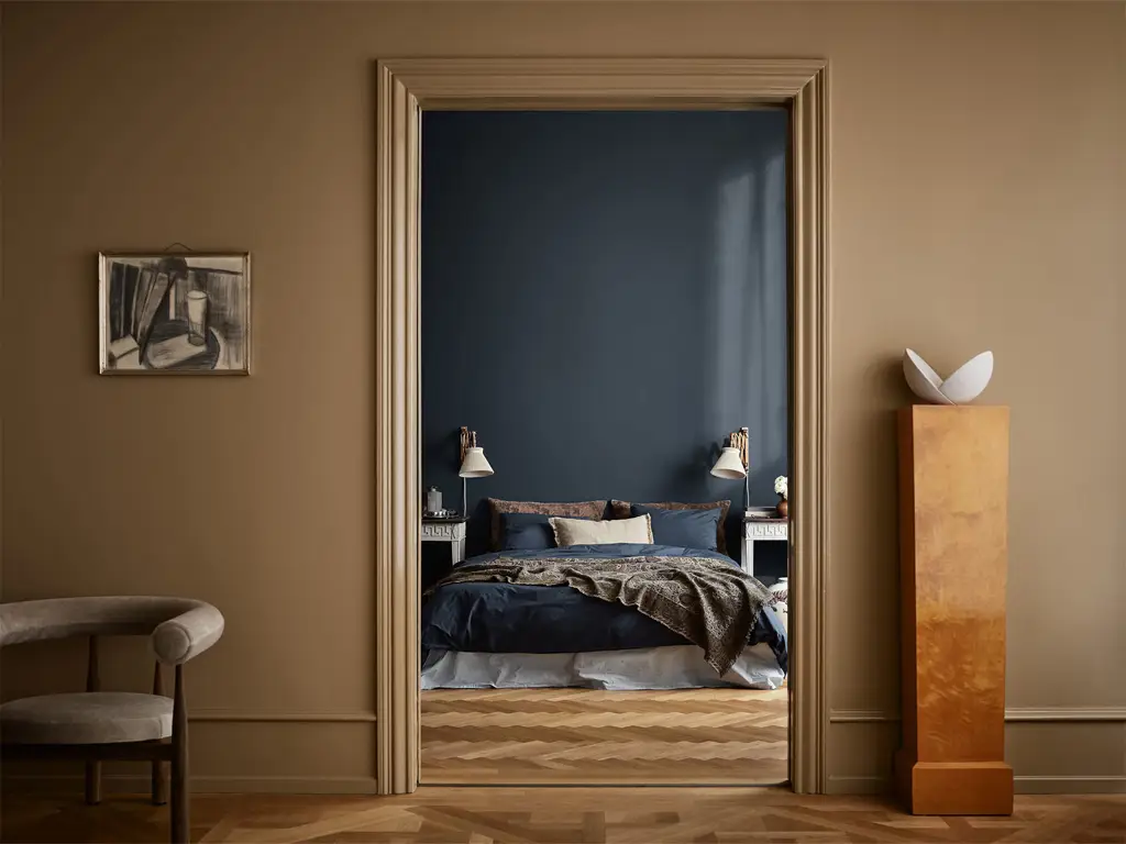 Elegant soverom sett gjennom døråpning, med mørk blå vegg malt i fargen Jotun Midnight, oppredd seng, nattbord. Fargen i fremre rom er Jotun Spice Brown