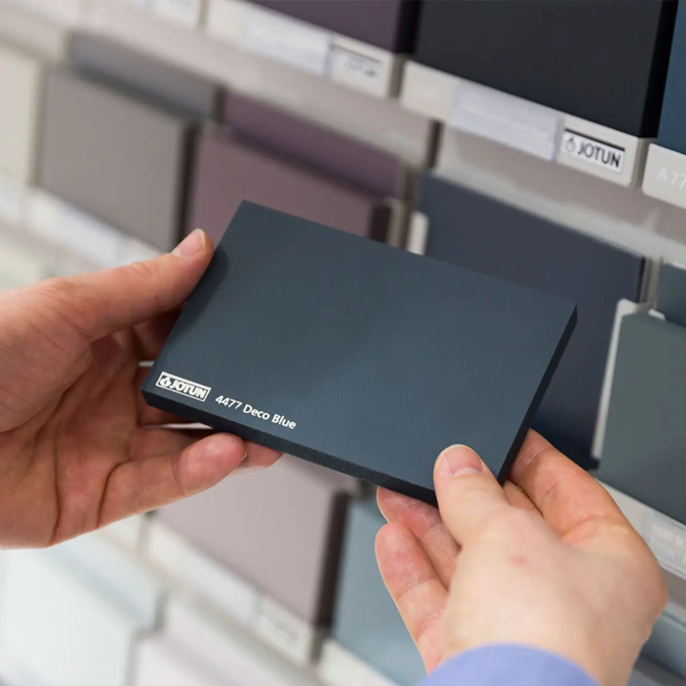 Person holder en fargeprøve fra Jotun med navnet "4477 Deco Blue" foran et utvalg av malingsprøver